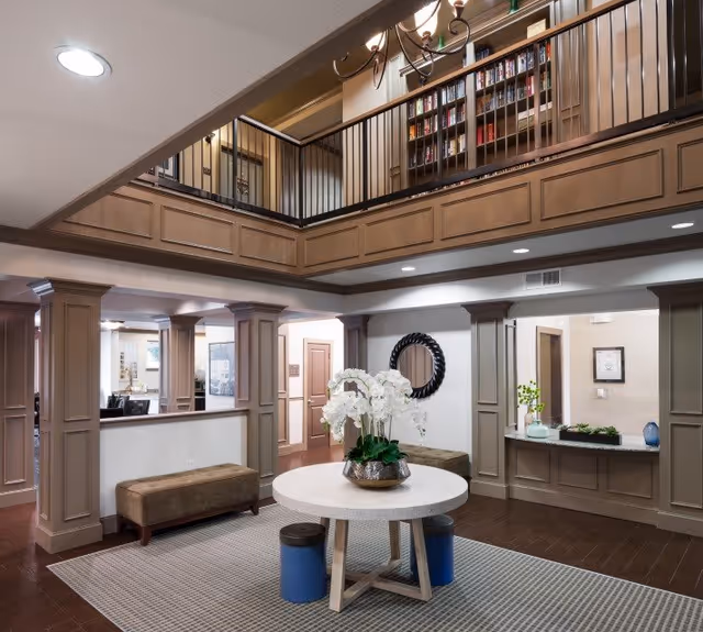 Interior view of a senior living facility lobby with a round table in the center holding a vase of white orchids. The room features beige walls with decorative columns, a carpeted area, and a wooden floor. There is a seating bench against the wall and a circular mirror hanging on the wall. Above, there is a second-floor balcony with a bookshelf filled with books and a chandelier hanging from the ceiling.