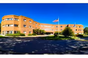 Wide exterior view of a three-story brick health care center building with an American flag on a flagpole in front, surrounded by greenery and a clear blue sky.
