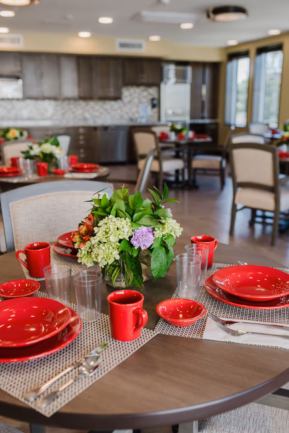 A dining table set with red plates, bowls, and mugs, clear drinking glasses, silverware, and a centerpiece of mixed flowers and green leaves in a vase. The background shows more tables and chairs in a dining area with a kitchen featuring dark cabinets and a tiled backsplash.