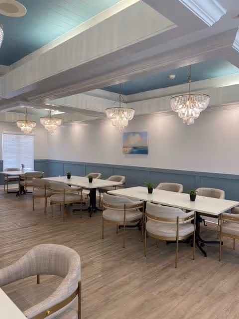 A bright and spacious dining room with multiple rectangular white tables and beige cushioned chairs arranged neatly. The room features light wood flooring, blue wainscoting on the walls, and a light blue ceiling with white beams. Elegant chandeliers hang from the ceiling, and small potted plants are placed on the tables. A painting of a seascape is mounted on the wall.