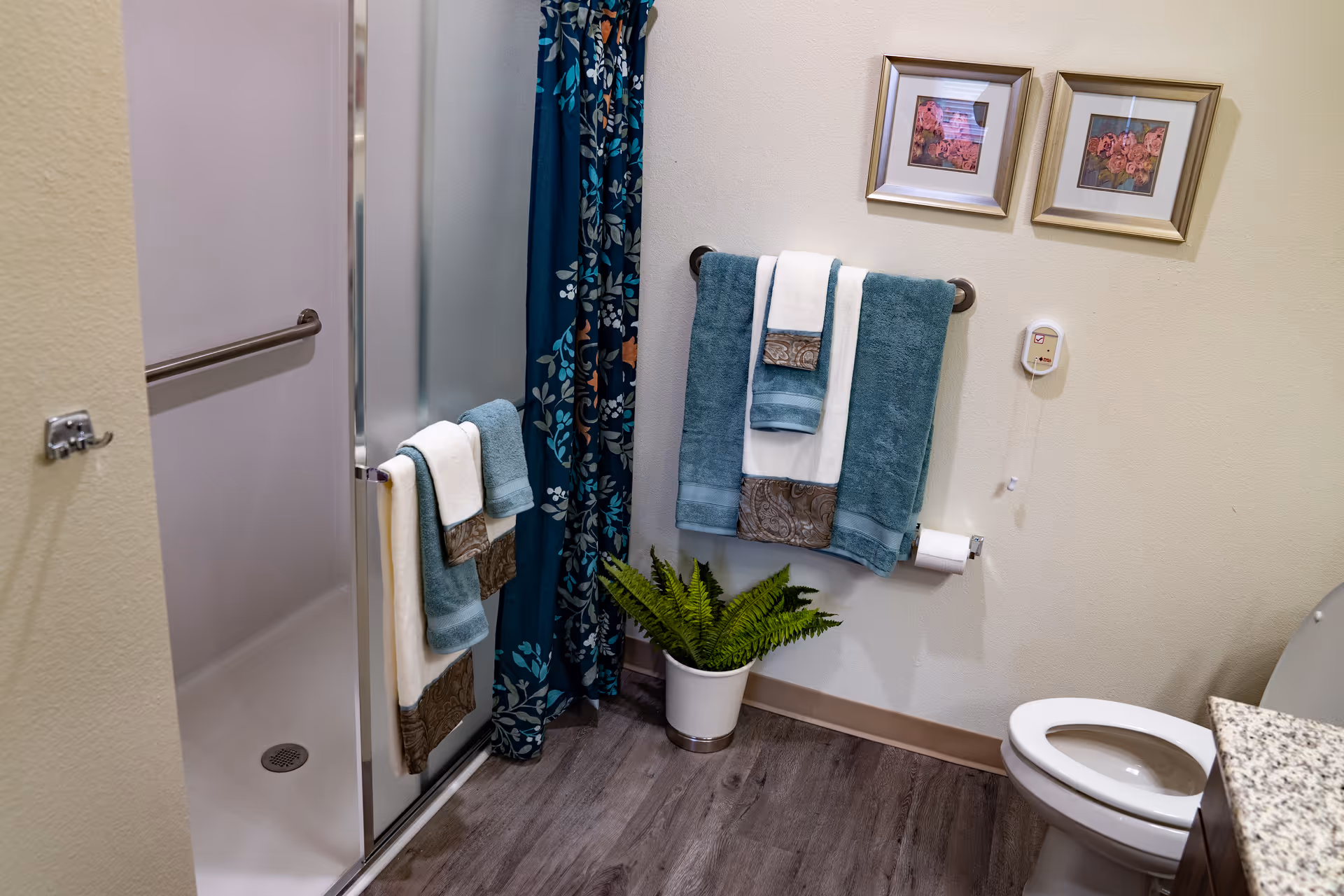A bathroom with a shower that has a frosted glass door and a blue floral shower curtain. There are multiple towels in teal and white colors hanging on towel racks. A potted green plant is placed on the floor near the wall. Two framed floral pictures hang above the towel rack. A toilet and part of a granite countertop are visible on the right side.