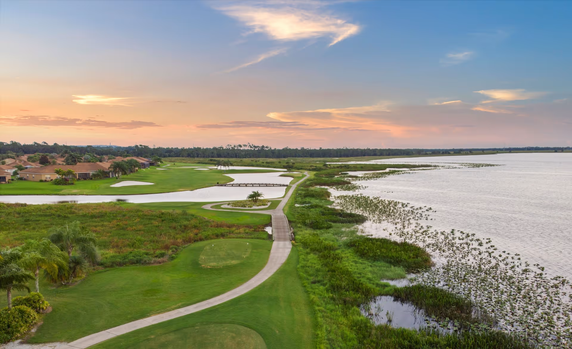 A scenic view of a golf course adjacent to a large body of water at sunset, with houses lining the left side and a pathway running through the green landscape.