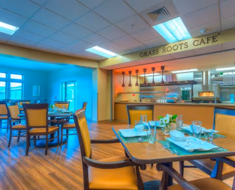 Interior view of a dining area named Grass Roots Cafe with wooden tables and chairs set with plates, glasses, and napkins. The room has a warm color scheme with wood flooring and ceiling lights. The kitchen area is visible in the background with hanging lights and cooking equipment.