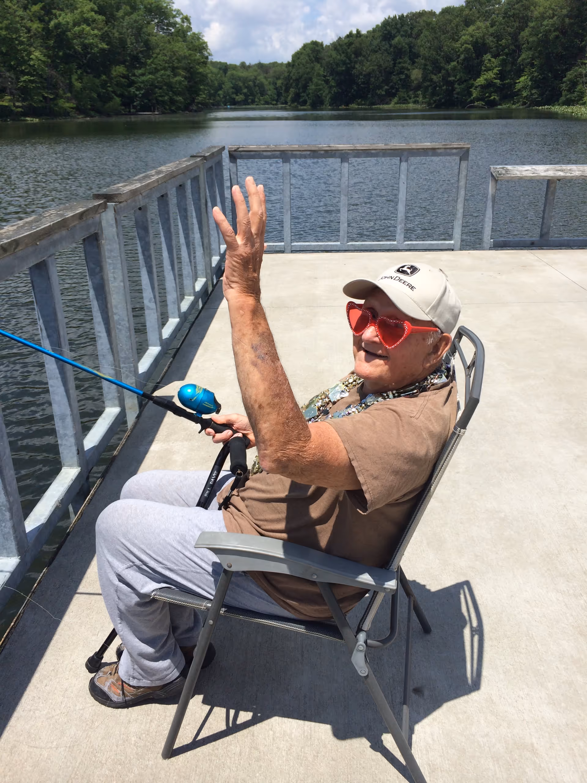 An elderly man wearing a beige John Deere cap and red heart-shaped sunglasses is sitting on a metal chair on a concrete dock by a lake. He is holding a fishing rod and waving with his other hand. The background shows calm water and green trees under a partly cloudy sky.