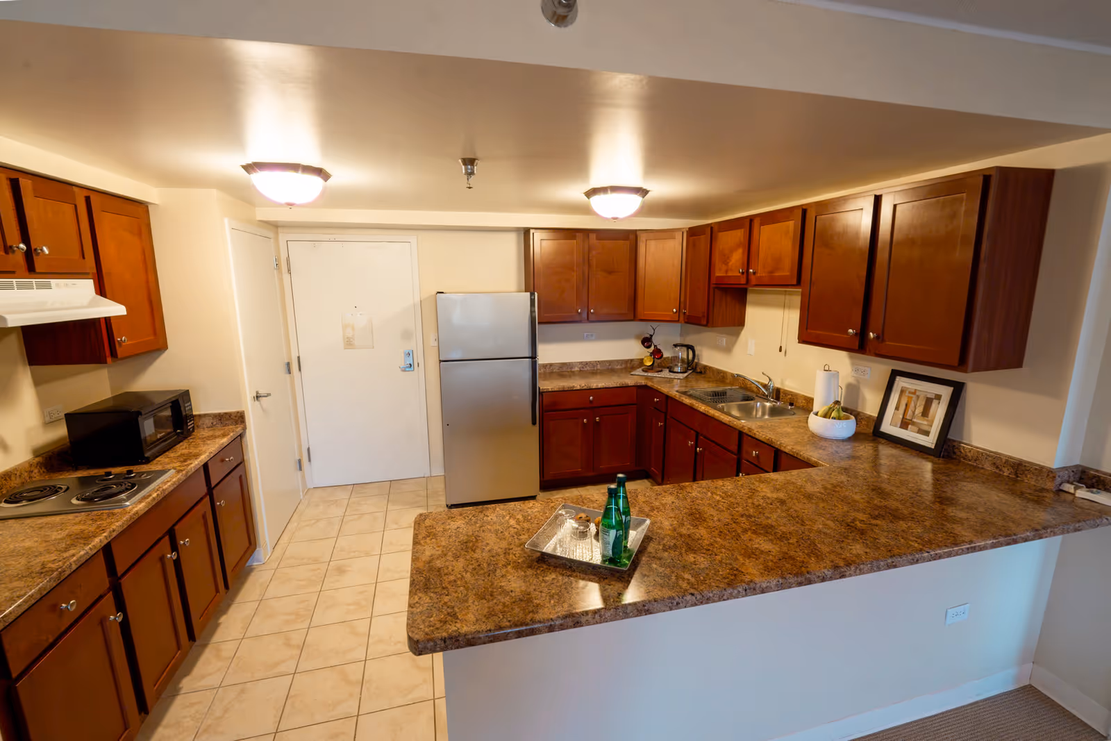 A kitchen area with brown wooden cabinets, a stainless steel refrigerator, a microwave, an electric stove, and a double sink. The countertops are a brown marble pattern, and there are two ceiling lights. On the counter, there is a tray with two green glass bottles and a glass, a bowl with bananas, and a framed picture leaning against the wall.