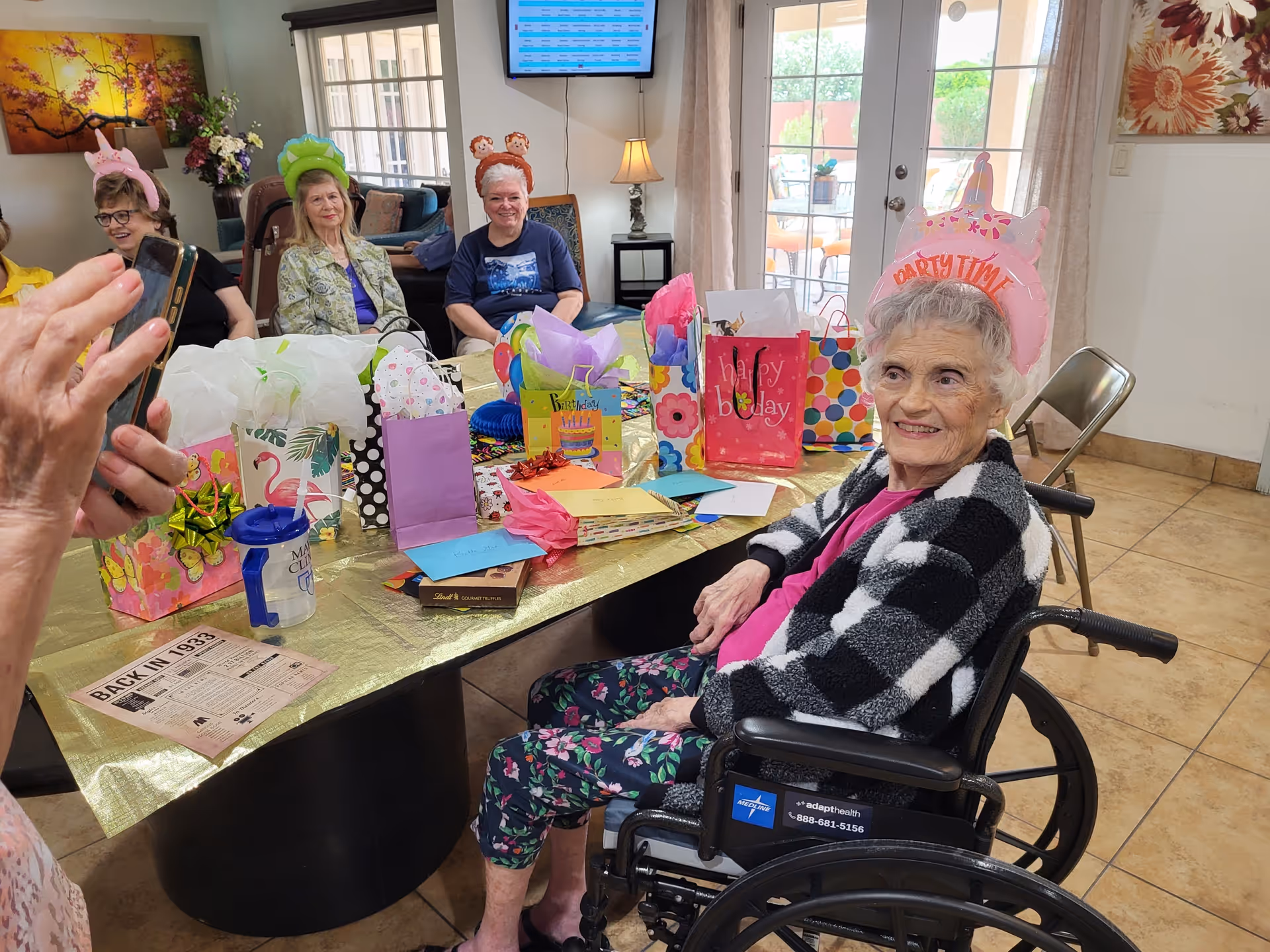 A group of elderly women sitting around a table decorated with colorful gift bags and birthday cards. One woman in a wheelchair is wearing a pink party hat that says 'Party Time' and smiling. Other women are wearing festive headbands and sitting in a cozy room with large windows and a TV mounted on the wall.
