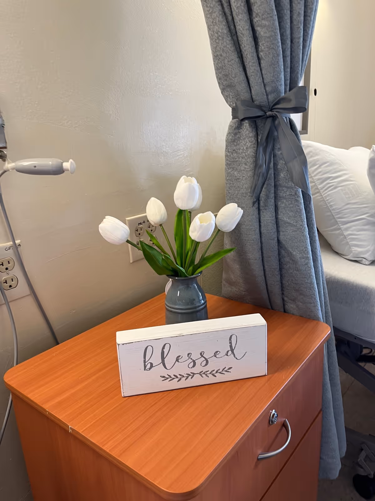 A wooden bedside table with a small vase holding white tulips and a decorative sign that reads 'blessed'. Next to the table is a hospital-style bed with white pillows and a gray privacy curtain tied back with a gray ribbon.