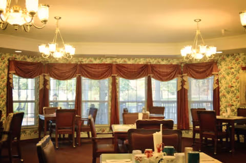 Dining room with multiple wooden tables and chairs arranged neatly. Large windows with red drapes and valances allow natural light to enter. The walls are decorated with floral wallpaper, and chandeliers hang from the ceiling providing warm lighting.