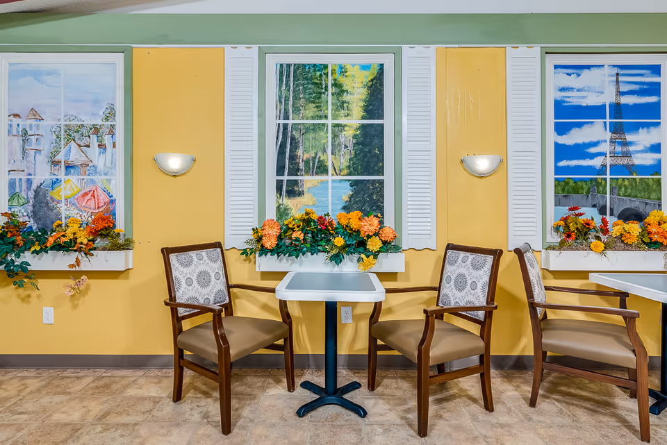 Small dining nook with two tables and wooden chairs against a yellow wall featuring three painted faux windows with flower boxes.