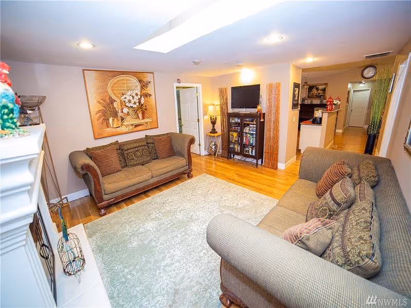 A cozy living room with two upholstered sofas facing each other on a light patterned area rug. The room features hardwood floors, a large framed artwork on the wall, a small table with a lamp, and a TV mounted on a cabinet. The space is well-lit with ceiling lights and a skylight, and a hallway leads to other parts of the facility.
