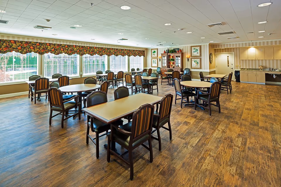 A spacious dining room with multiple tables and chairs arranged neatly. Large windows with floral valances allow natural light to fill the room. The walls are decorated with framed artwork and a wooden cabinet. The floor has a wood-like finish, and there is a countertop area with cabinets along one wall.