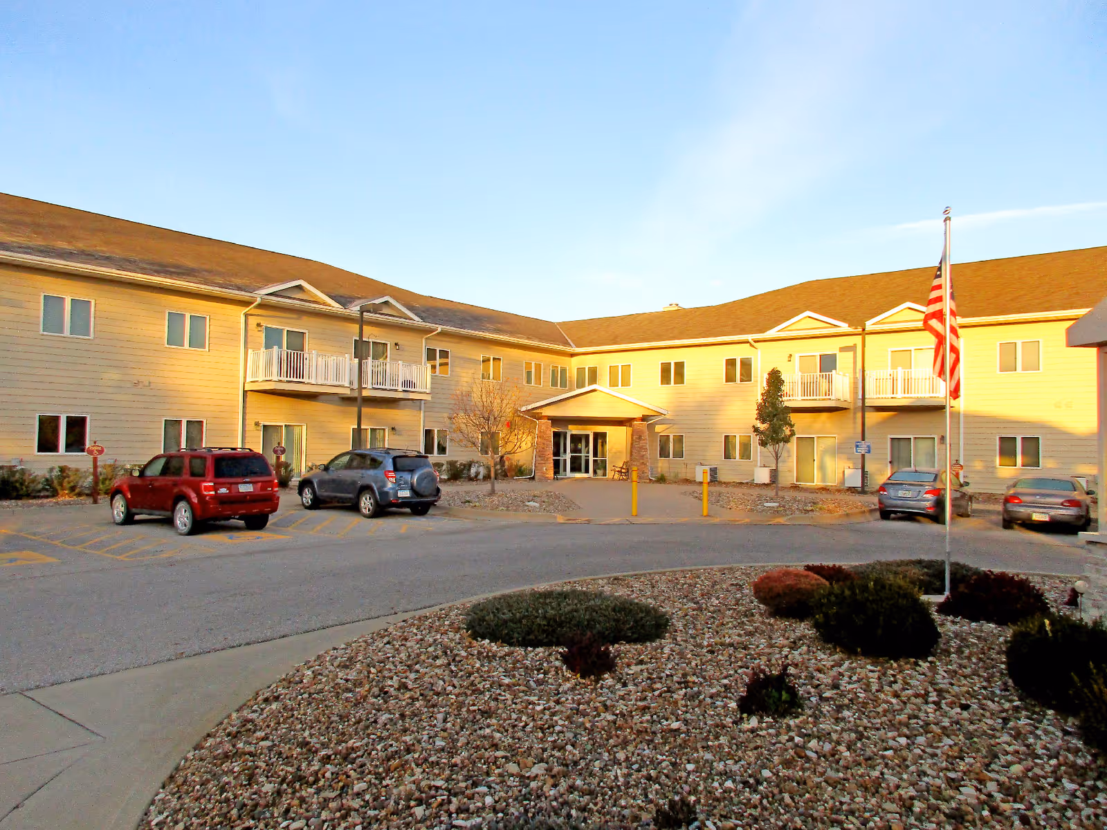 Front exterior of a two-story senior living building with parked cars, an American flag, and a rock garden in the foreground.
