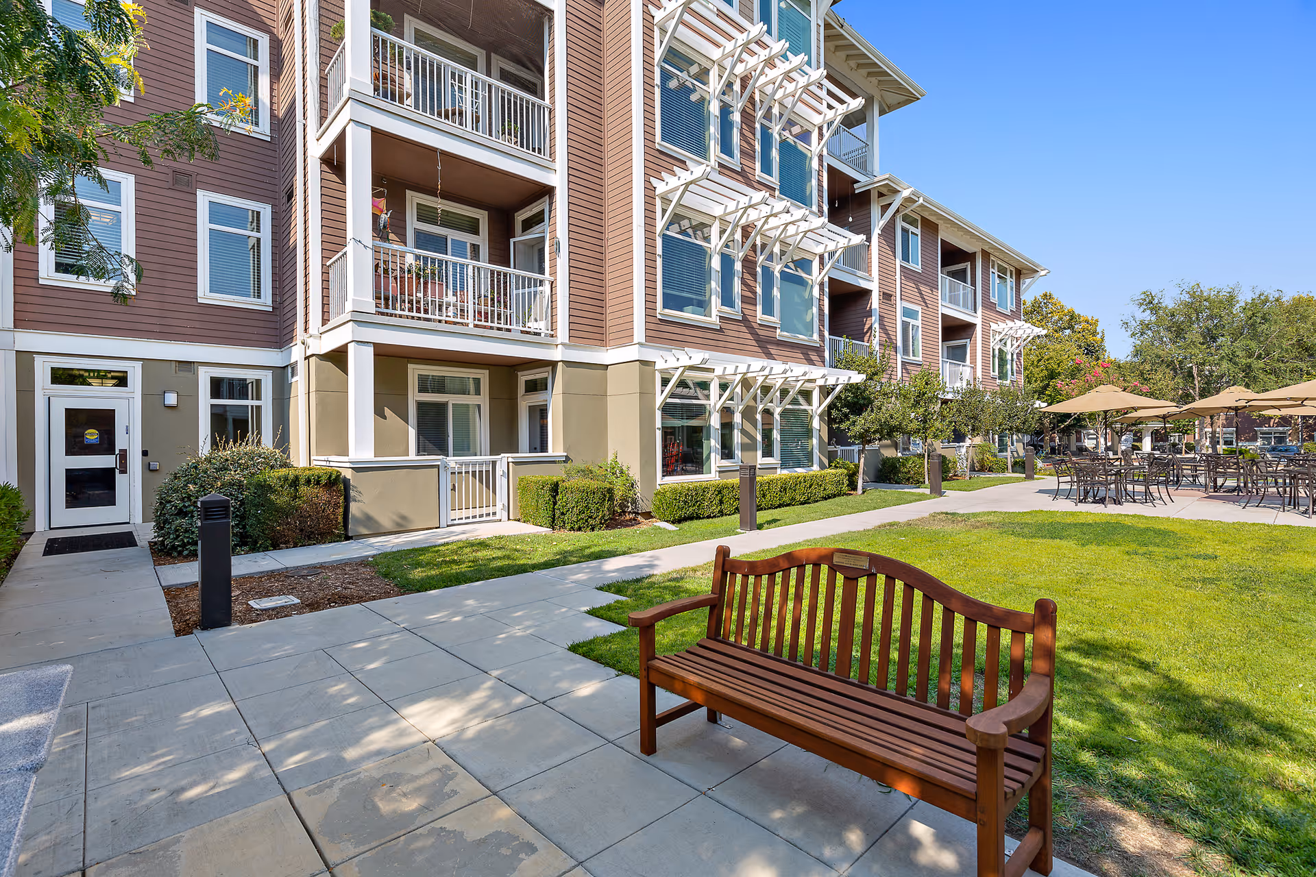 Outdoor courtyard of a multi-story senior living building with balconies, a wooden bench, lawn, and patio seating with umbrellas.
