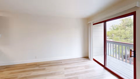 Empty living room with light wood floors and a sliding glass door opening onto a balcony.