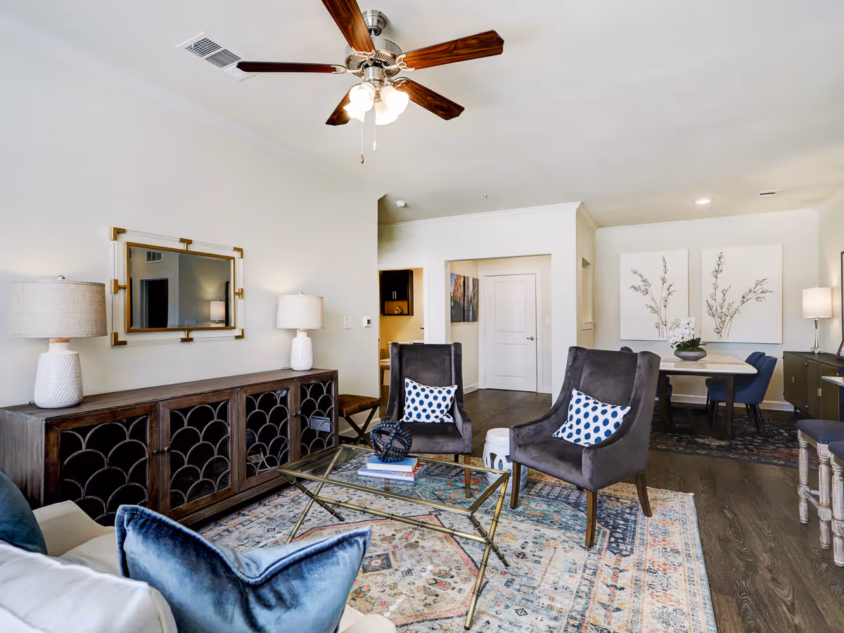 A bright and spacious living room with a ceiling fan, two dark gray armchairs with patterned pillows, a glass coffee table on a colorful patterned rug, a wooden sideboard with two lamps and a mirror above it, and a dining area with a table, chairs, and floral artwork on the wall.