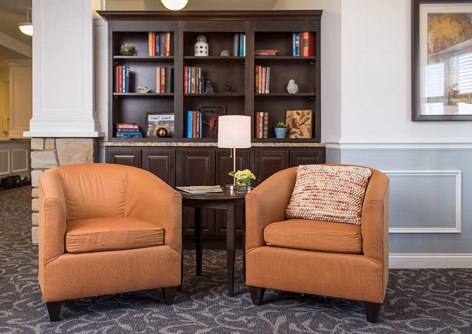 Two orange upholstered armchairs with a small round wooden table between them, holding a lamp and a small flower arrangement. Behind the chairs is a dark wooden bookshelf filled with books and decorative items. The room has a patterned carpet and light-colored walls with molding and a framed picture on the right side.