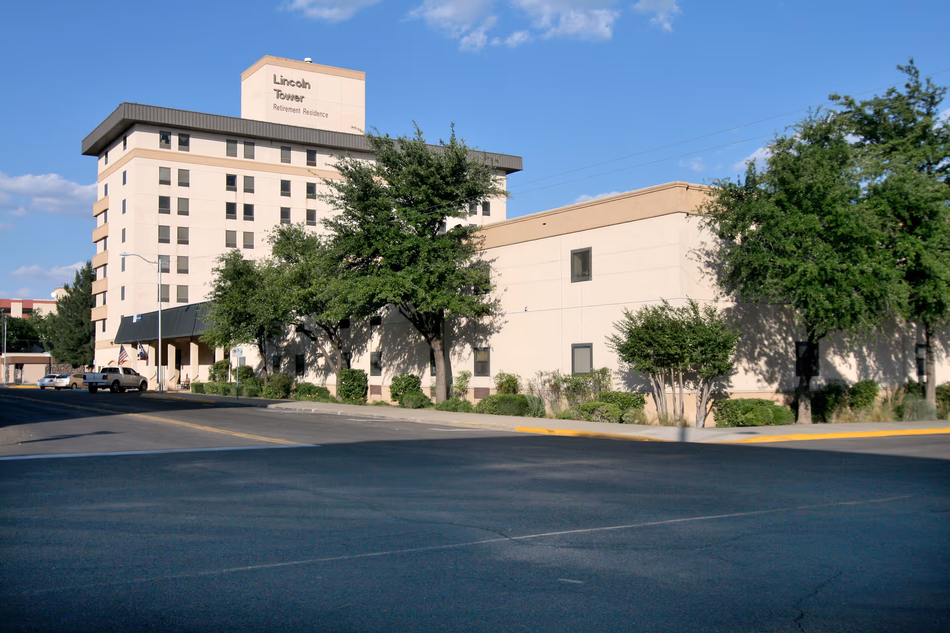 Exterior view of a multi-story beige building labeled Lincoln Tower Retirement Residence with trees and parked cars along the street in front under a clear blue sky.