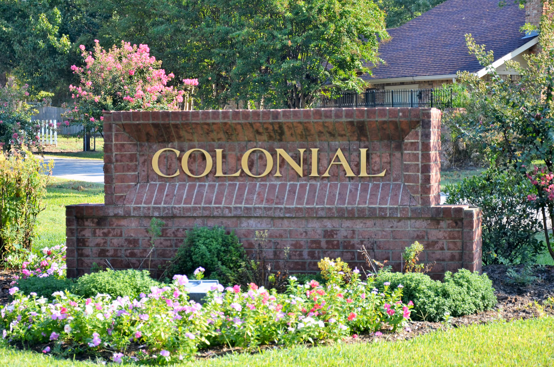 A brick sign with the word 'COLONIAL' in gold letters surrounded by green bushes and pink flowers, with trees and a building roof visible in the background.