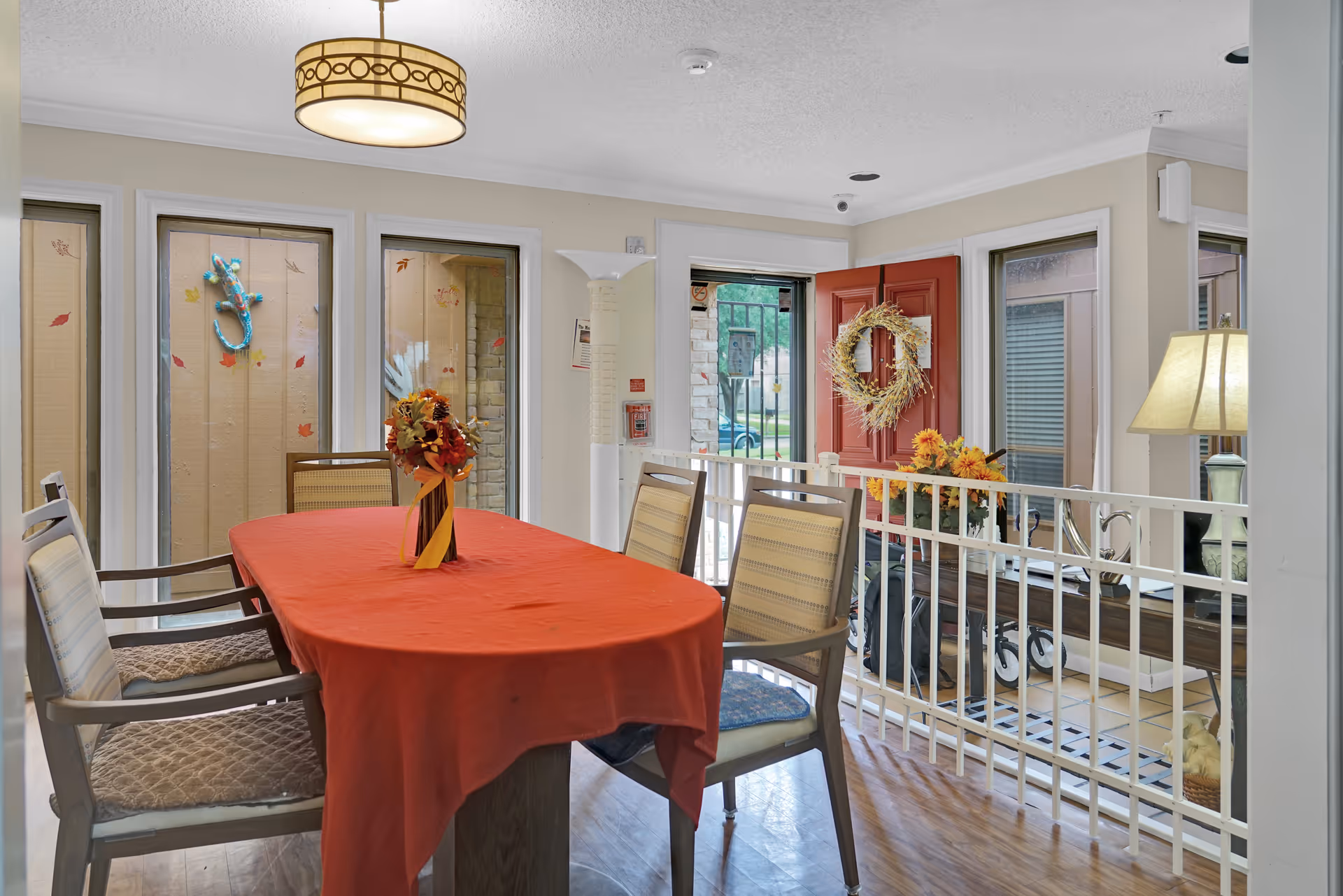 A dining area with a rectangular table covered with an orange tablecloth and six chairs around it. A vase with autumn-themed flowers is placed in the center of the table. The room has large windows with fall leaf decorations on the glass. A red door with a wreath is open, showing a glimpse of the outside. There is a white railing separating the dining area from a lower level with a lamp and more autumn decorations.