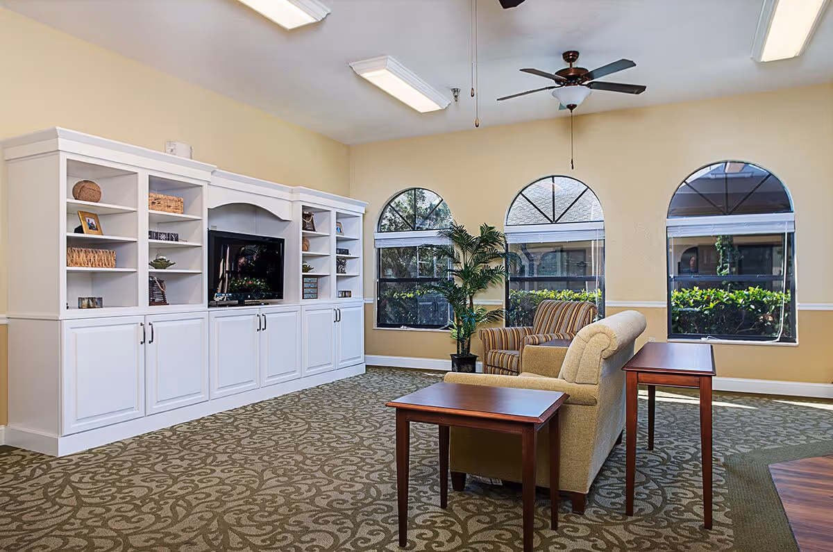 A cozy living room area with a beige couch and a striped armchair facing a large white entertainment center with a flat-screen TV. The room has three arched windows with views of greenery outside, a ceiling fan, and patterned carpet flooring.