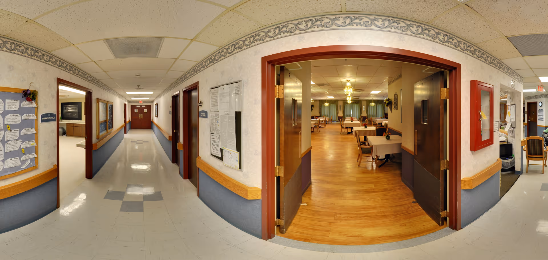 A corridor in a senior living facility opens through double doors into a dining room with tables and chairs set on a wood floor.