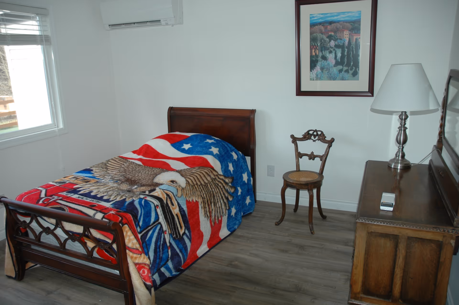 A small bedroom with a single wooden bed covered by a colorful blanket featuring an eagle and American flag design. Next to the bed is an antique wooden chair with a woven seat. A wooden dresser with a lamp and a small device on top is positioned against the wall, which also has a framed landscape painting. A window with blinds is on the left side of the room.