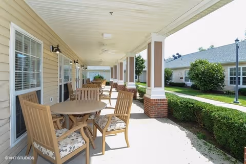 Covered outdoor patio area with round tables and wooden chairs with patterned cushions, adjacent to a beige building with windows and wall-mounted lights. The patio overlooks a landscaped garden with bushes, a walkway, and another building in the background under a clear sky.