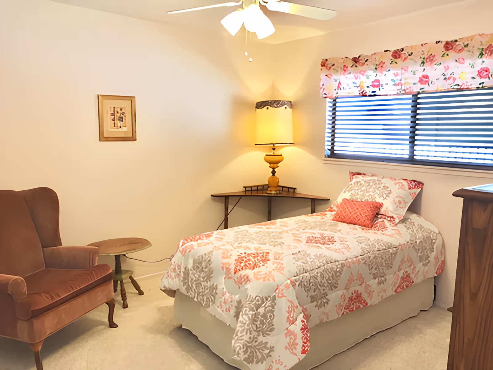 A cozy bedroom with a single bed covered in a patterned quilt with red, beige, and white colors. There is a small red pillow on the bed. Next to the bed is a corner table with a yellow lamp. A brown upholstered armchair and a small round wooden side table are positioned against the wall. A window with horizontal blinds and a floral valance is above the bed. The room has beige carpet and light-colored walls with a ceiling fan overhead.