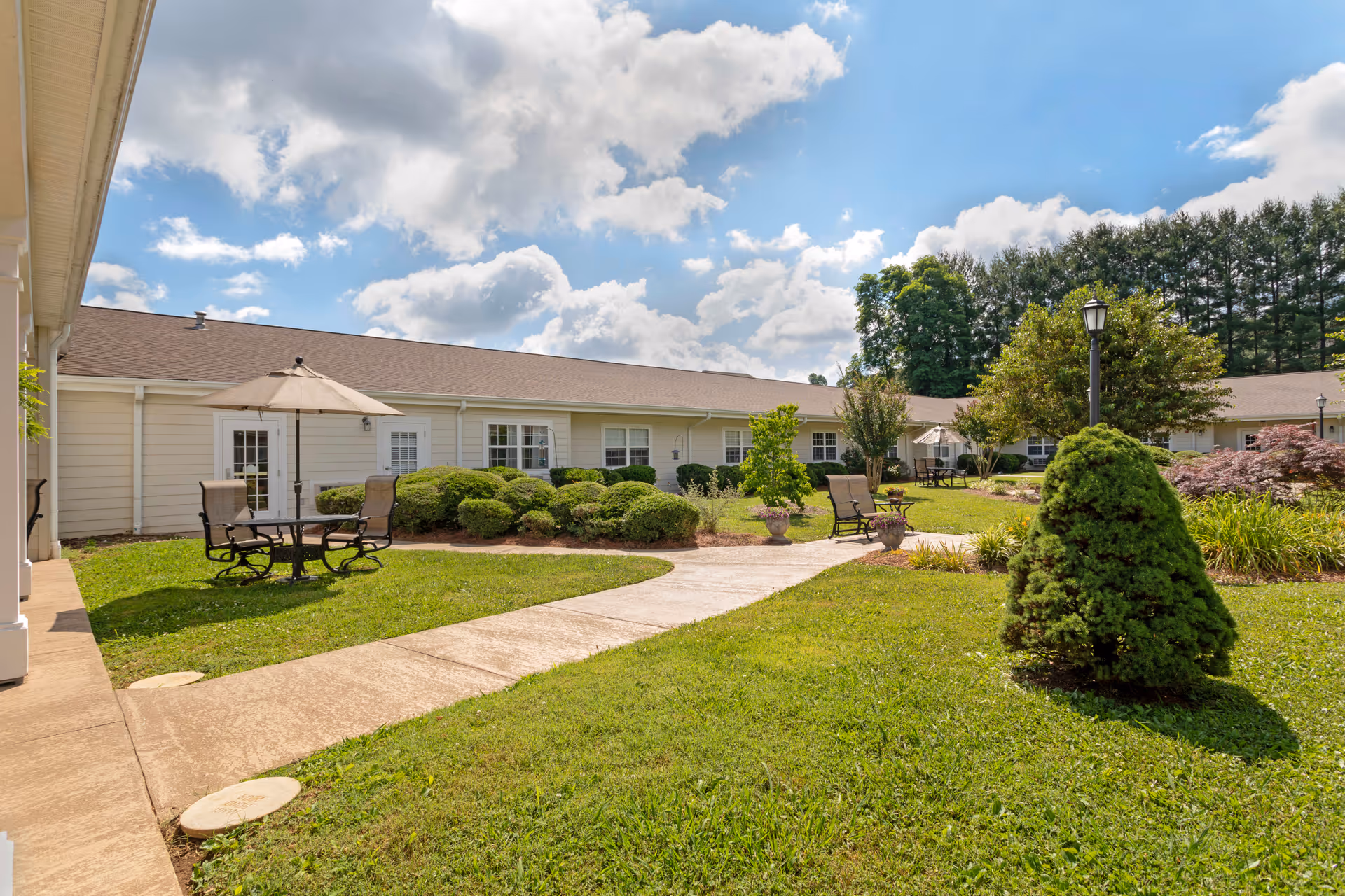 Outdoor courtyard area at Brookdale Johnson City featuring a paved walkway, green grass, trimmed bushes, small trees, and outdoor seating with tables and chairs under umbrellas. The building with beige siding and multiple windows surrounds the courtyard under a partly cloudy blue sky.