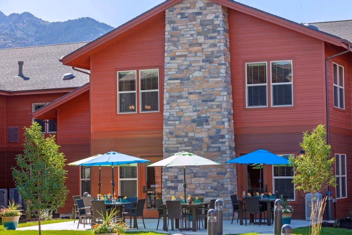 Outdoor patio area at The Lodge in North Ogden with tables and chairs under blue and white umbrellas, surrounded by small trees and plants, with a red building and stone chimney in the background and mountains visible behind.