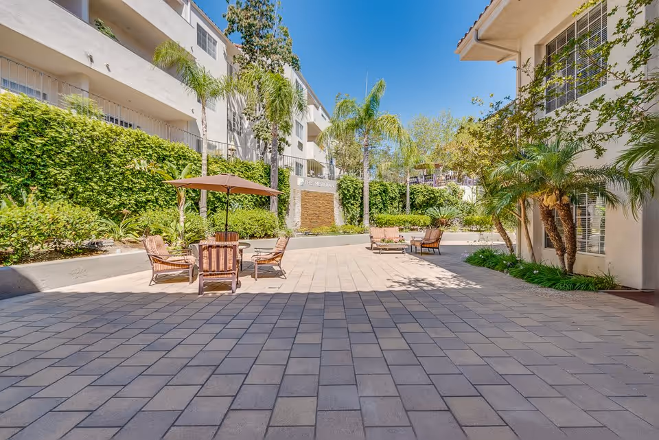 Outdoor courtyard area at Meridian at Anaheim Hills featuring a paved patio with several chairs and tables, some shaded by umbrellas. The space is surrounded by greenery including palm trees, bushes, and other plants, with multi-story residential buildings on either side under a clear blue sky.