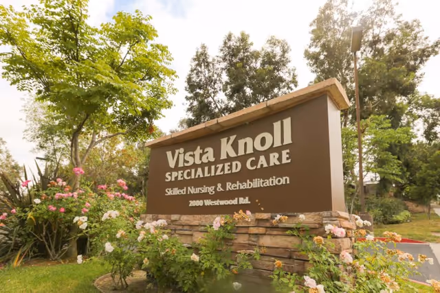 Outdoor view of a large brown and stone sign for Vista Knoll Specialized Care, Skilled Nursing & Rehabilitation, located at 2000 Westwood Rd, surrounded by green trees and flowering plants.