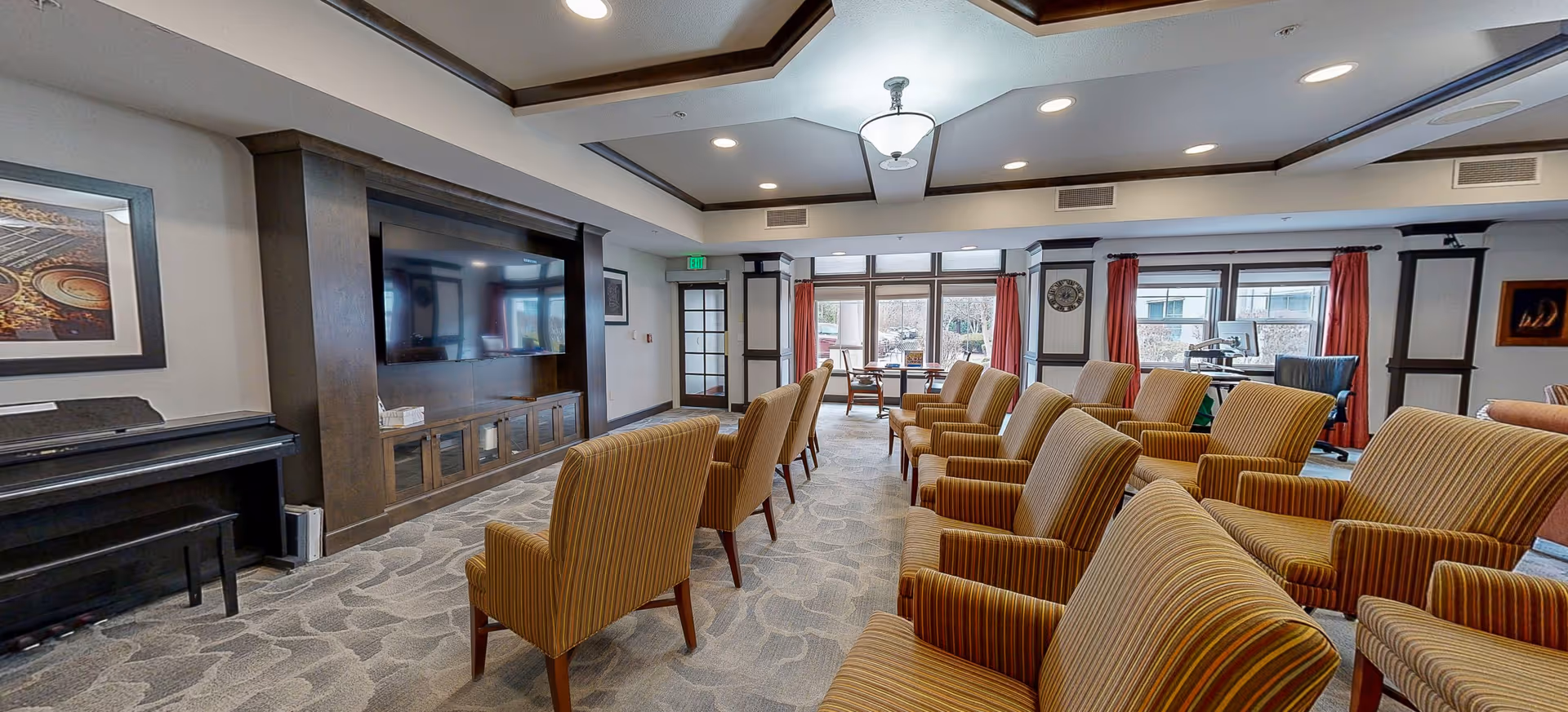 A spacious common area with multiple striped armchairs arranged in rows facing a large flat-screen TV mounted on a dark wooden entertainment center. The room has a patterned carpet, a piano on the left side, large windows with red curtains letting in natural light, and a ceiling light fixture.