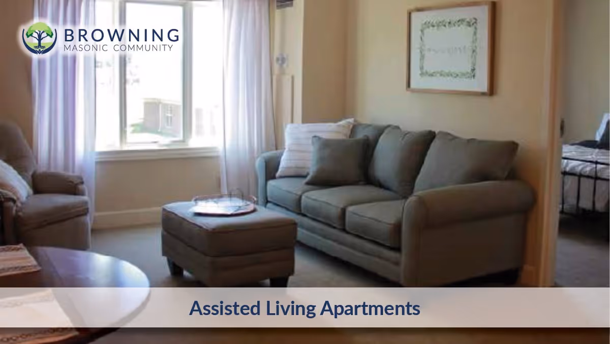 A cozy living room in an assisted living apartment featuring a gray sofa with cushions, a matching ottoman, an armchair, a coffee table, and a window with white curtains letting in natural light. A framed picture hangs on the wall, and a bedroom is partially visible through an open doorway.
