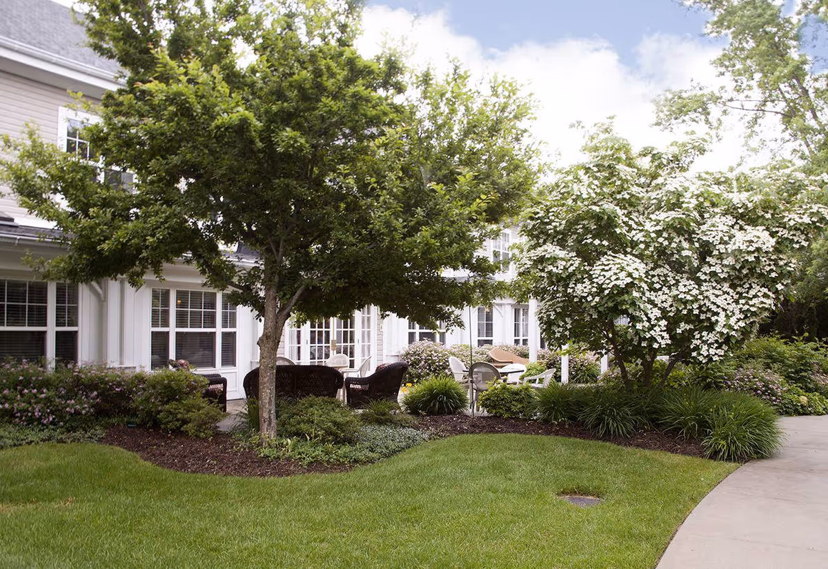 Outdoor garden area at Sunrise of Holbrook with green grass, trees, flowering bushes, and patio furniture including chairs and tables near the building.