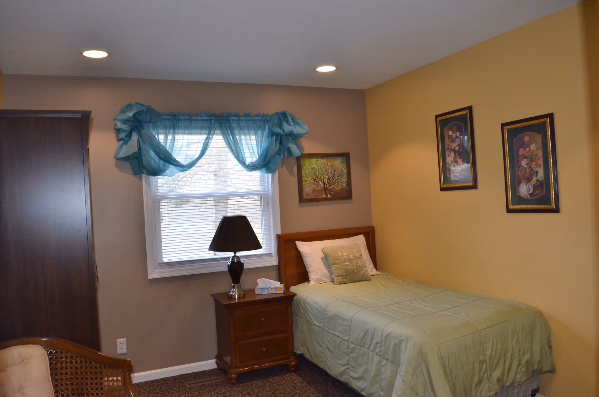 A small bedroom with a single bed covered in a light green comforter and a matching pillow. Next to the bed is a wooden nightstand with a black lamp and a box of tissues. The room has two-tone walls, one side painted beige and the other yellow. There are three framed floral paintings on the walls and a window with blue sheer curtains above the nightstand. A wooden wardrobe and a wicker chair with a cushion are partially visible.