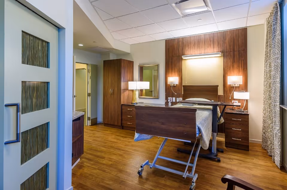 A well-lit senior living facility bedroom with a hospital-style adjustable bed, wooden furniture including a wardrobe, dresser, and nightstands, lamps on each side of the bed, a large window with patterned curtains, and a door leading to an adjacent bathroom.