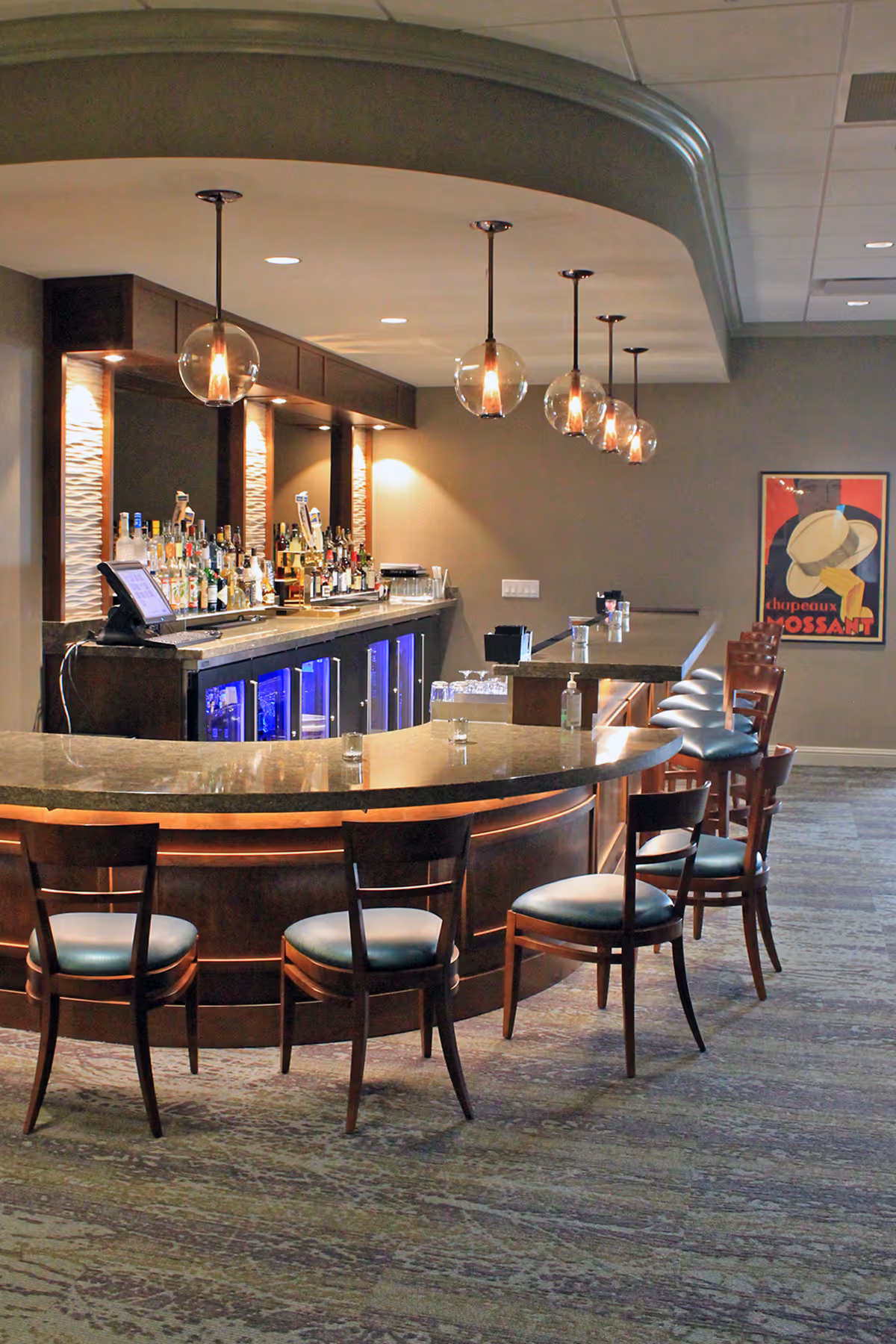Interior view of a bar area with a curved counter, several wooden chairs with blue cushions, hanging globe pendant lights, and a variety of bottles and glasses behind the counter. A framed poster is visible on the wall in the background.