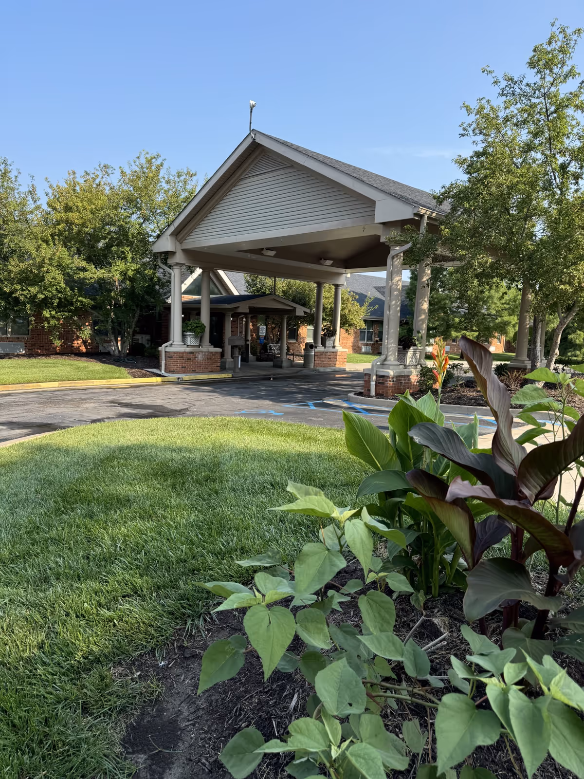Entrance of The Bluffs facility with a covered drop-off area supported by columns. The building is surrounded by green grass, trees, and various plants under a clear blue sky.