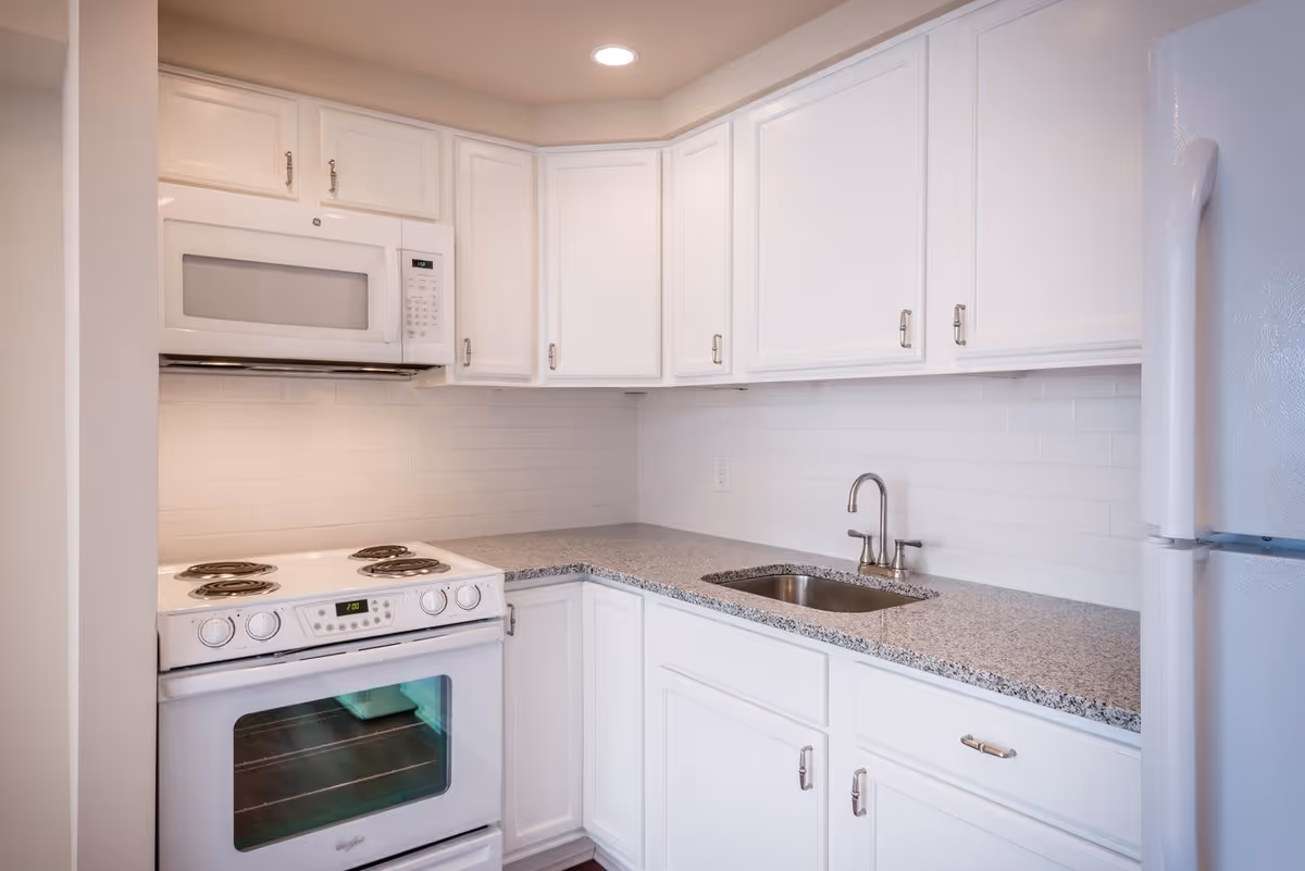 A clean, modern kitchen with white cabinets, a white electric stove with four burners, a white microwave above the stove, a stainless steel sink with a faucet, and a white refrigerator. The countertop is gray granite and the backsplash is white subway tile.