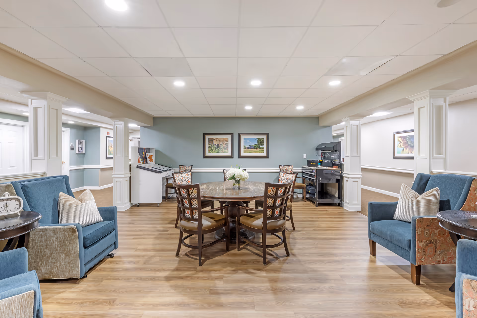 A well-lit common area with a round wooden table surrounded by six chairs with patterned cushions. Two blue armchairs with beige pillows are positioned on either side of the room, each next to a small round wooden table. The walls are painted light blue and beige with white trim, and two framed pictures hang on the far wall. The floor is wood laminate, and the ceiling has recessed lighting. There is a white cabinet and a black cart with equipment against the back wall.