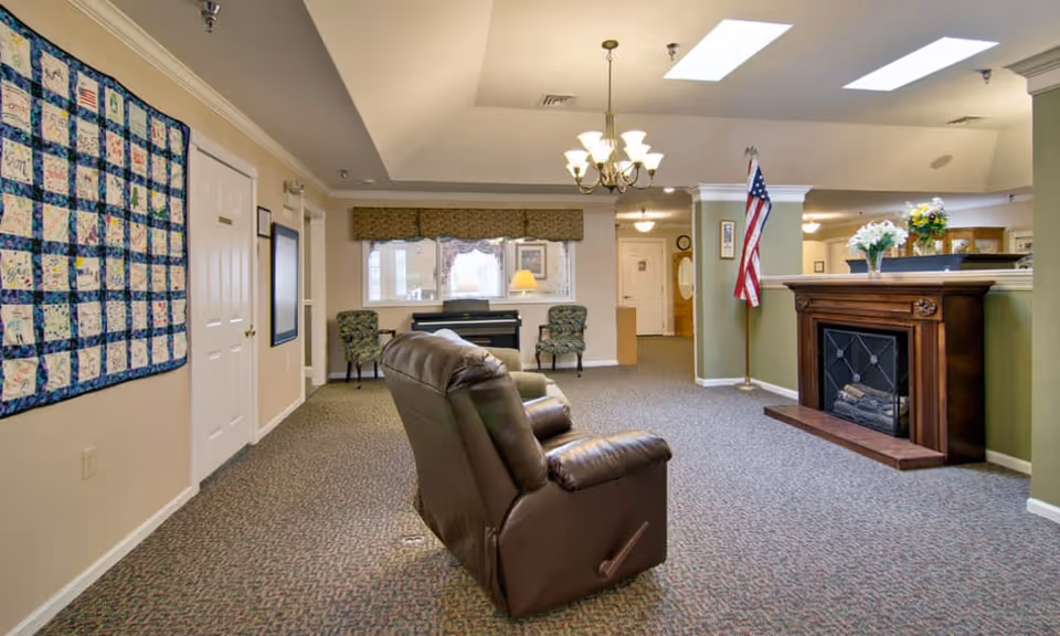 A spacious living room area in a senior living facility with a brown leather recliner in the center, a wooden fireplace with flowers on top, an American flag on a stand, two patterned chairs near a window with a piano between them, and a quilt hanging on the wall.