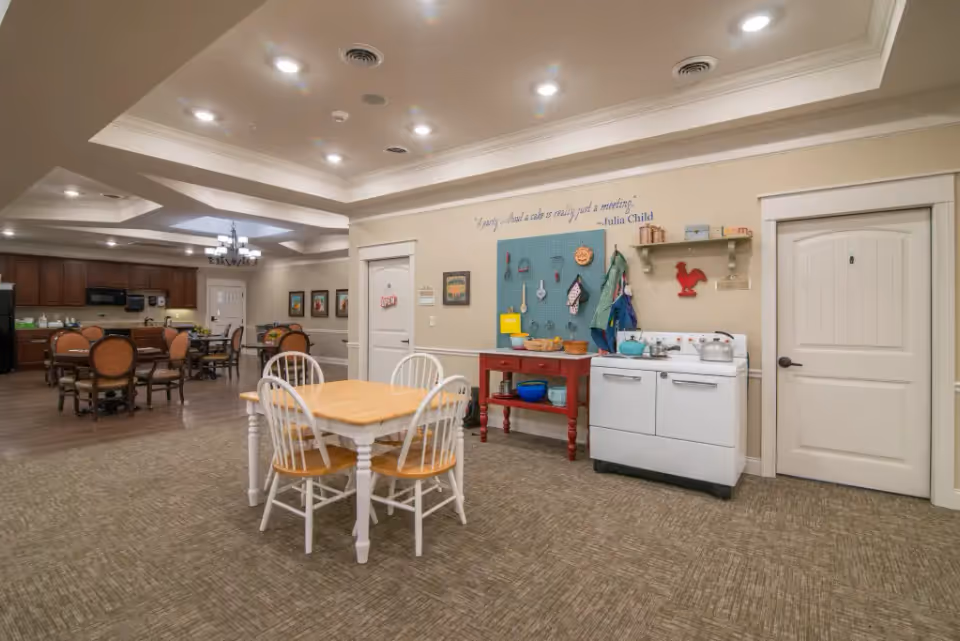 Spacious communal dining and activity room with a wooden table and chairs in the foreground, dining tables in the background, and a small kitchen/craft station along the wall.