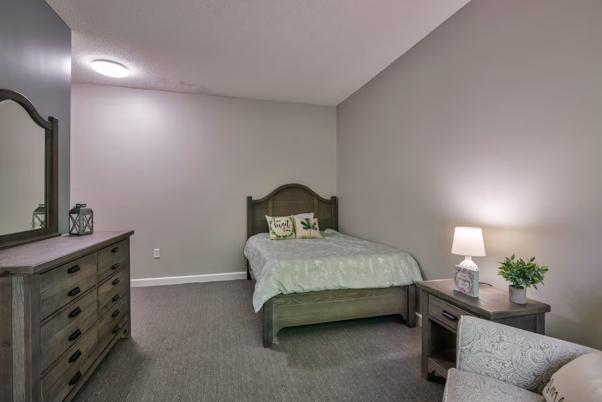 Furnished bedroom with a wooden bed, dresser, nightstand with a lamp and plant, and an upholstered chair.