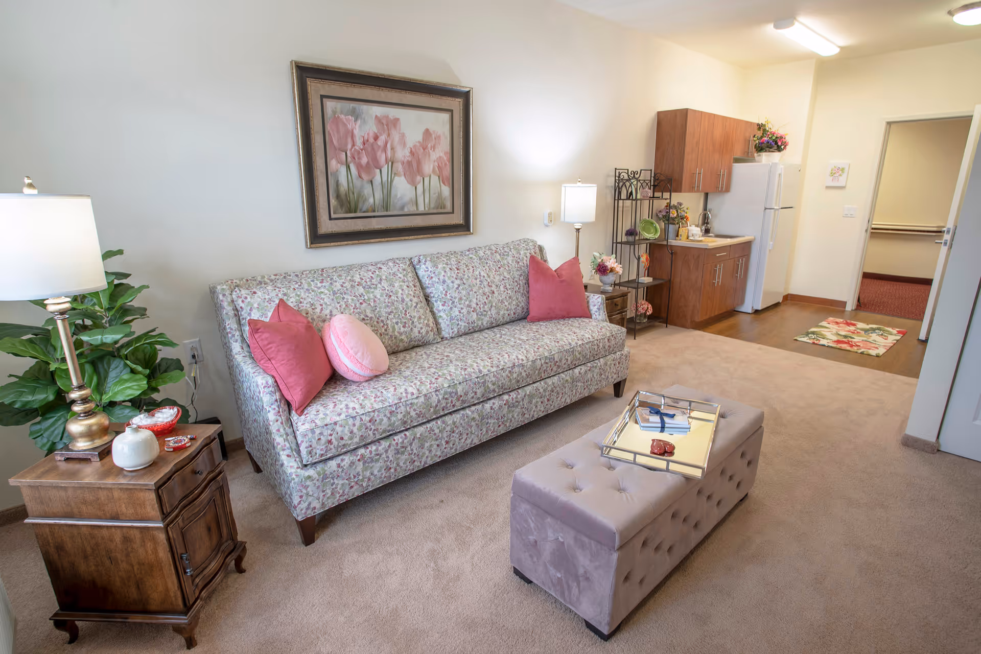 Cozy living room with a floral upholstered sofa, pink pillows, a tufted ottoman, side table and a small kitchenette in the background.