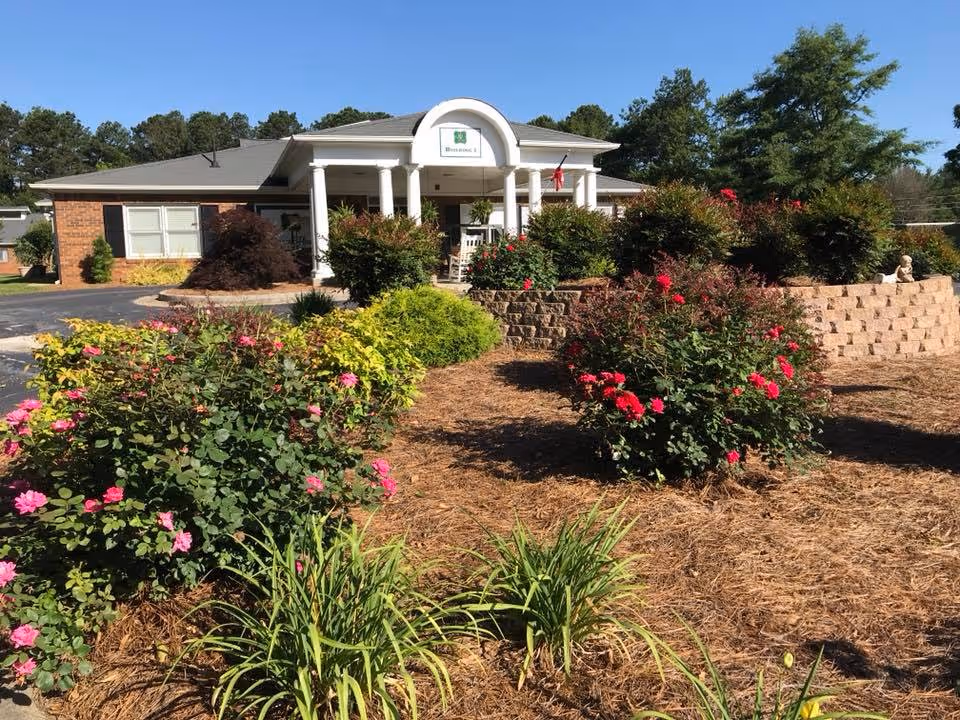 Front of a single-story brick building with a white columned entrance and flowering shrubs in the landscaped foreground.