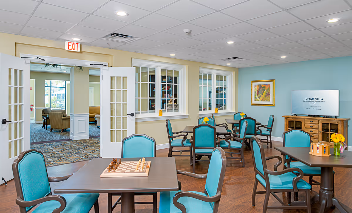 Bright senior living common room with teal chairs and tables, a chessboard on a table, a TV, and open double doors to a lounge.