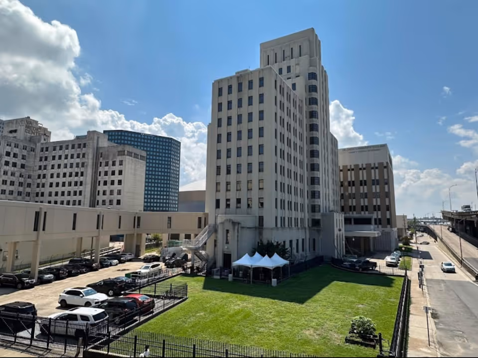Exterior view of a multi-story institutional building with a fenced lawn, parked cars, and a pedestrian skybridge under a blue sky.