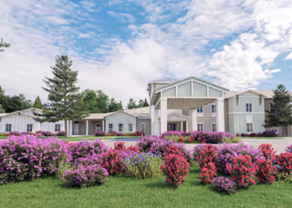 Exterior view of a senior living facility building with a covered entrance. The building is surrounded by green grass and vibrant purple and pink flowering bushes under a partly cloudy blue sky.
