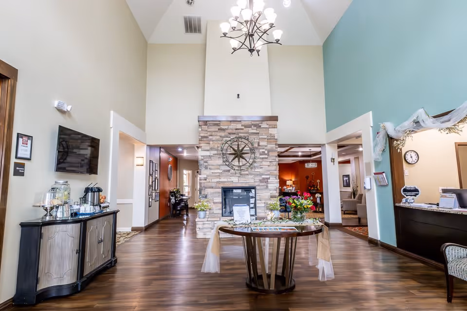 Interior view of a senior living facility lobby with a high ceiling and a stone fireplace in the center. A round table with flowers and informational materials is placed in front of the fireplace. To the left, there is a cabinet with a beverage dispenser and coffee setup. To the right, there is a reception desk with a clock on the wall behind it. The floor is wooden, and the walls are painted in light colors with some teal and beige accents.